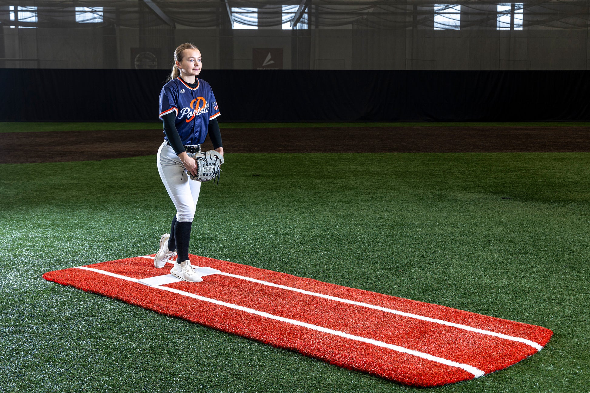 A softball player stands on a red pitching mound indoors, positioned on the Portolite Pro Spiked Game Mat with Lines / Lanes—built for collegiate and pro-level pitching—for optimal field maintenance, wearing a navy jersey and white pants, holding a glove and ball, ready to pitch.