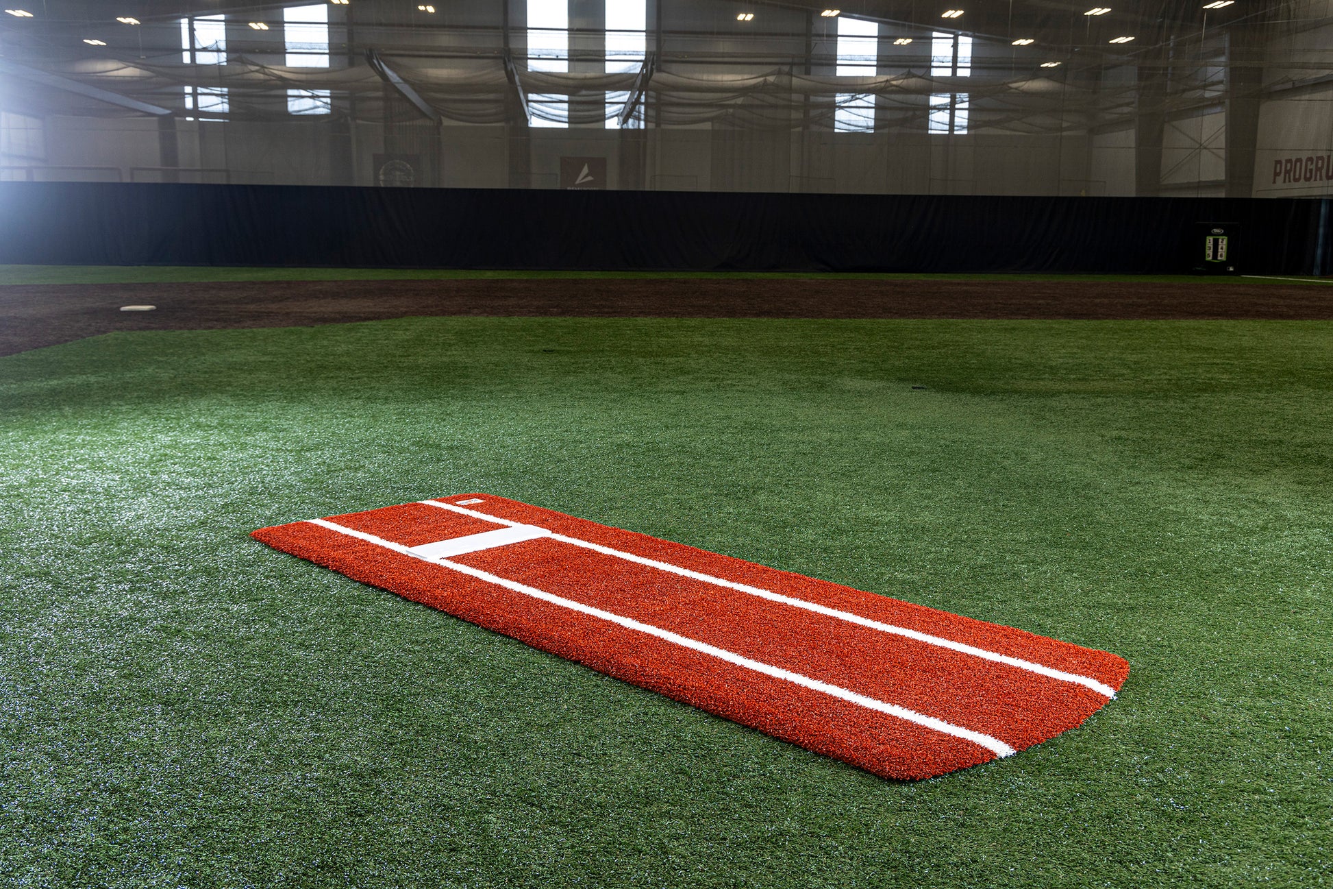 A Portolite Pro Spiked Game Mat with Lines / Lanes, designed for collegiate and pro-level pitching, sits on green turf inside an indoor baseball or softball training facility.