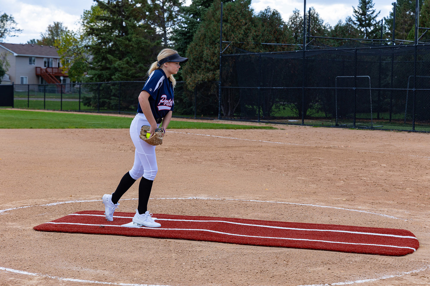 A softball pitcher in uniform stands on the mound atop the Portolite Pro Spiked Game Mat with Lines / Lanes, built for collegiate and pro-level pitching, holding a glove and ready to pitch on a sunny day with trees and houses visible in the background.