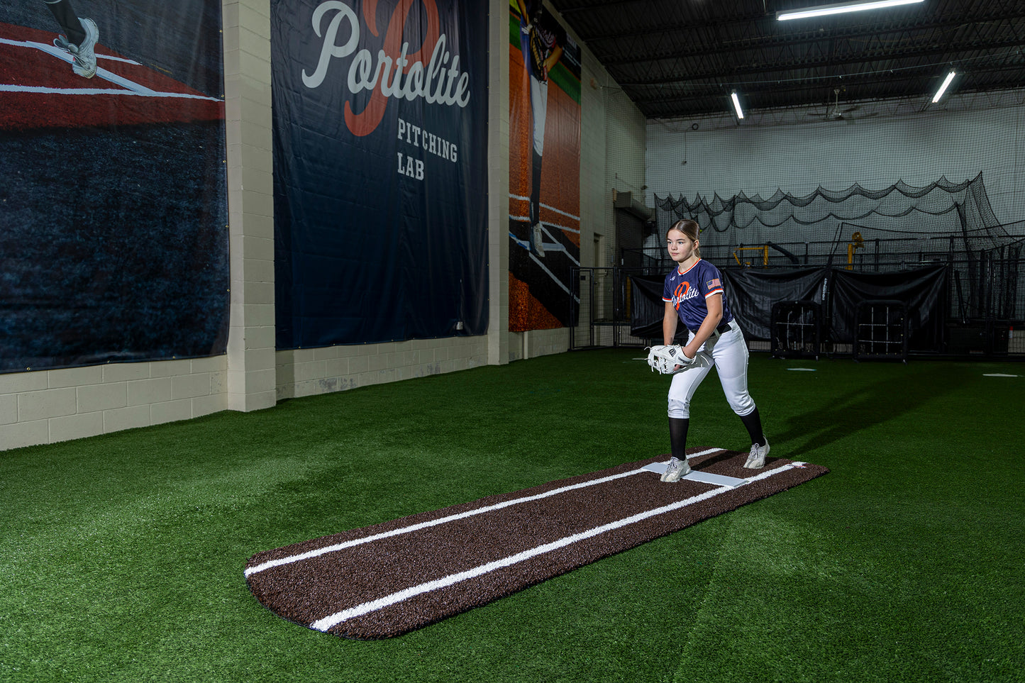 A softball player stands on the Portolite Signature Practice Mat with Spikes with Lines / Lanes, designed for 12U pitchers, preparing to pitch inside a facility labeled Portalite Pitching Lab.