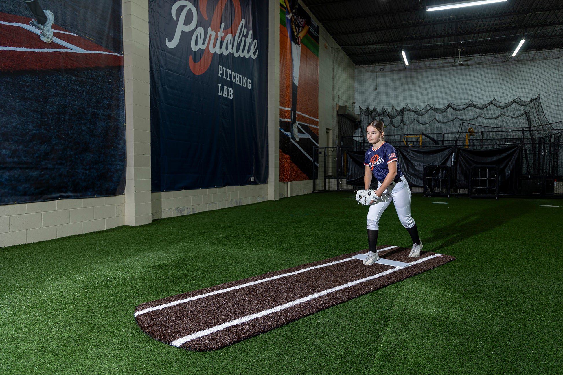 A softball player stands on the Portolite Signature Practice Mat with Spikes with Lines / Lanes, designed for 12U pitchers, preparing to pitch inside a facility labeled Portalite Pitching Lab.