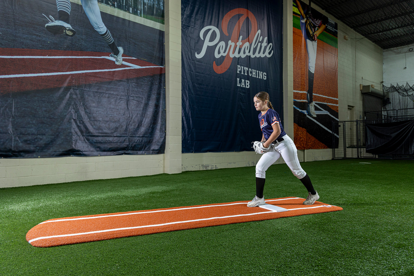 A softball player stands ready to pitch on a Portolite Signature Practice Mat with Spikes with Lines / Lanes, built for 12U pitchers, in an indoor facility with synthetic turf and a Portolite Pitching Lab banner displayed prominently behind her.