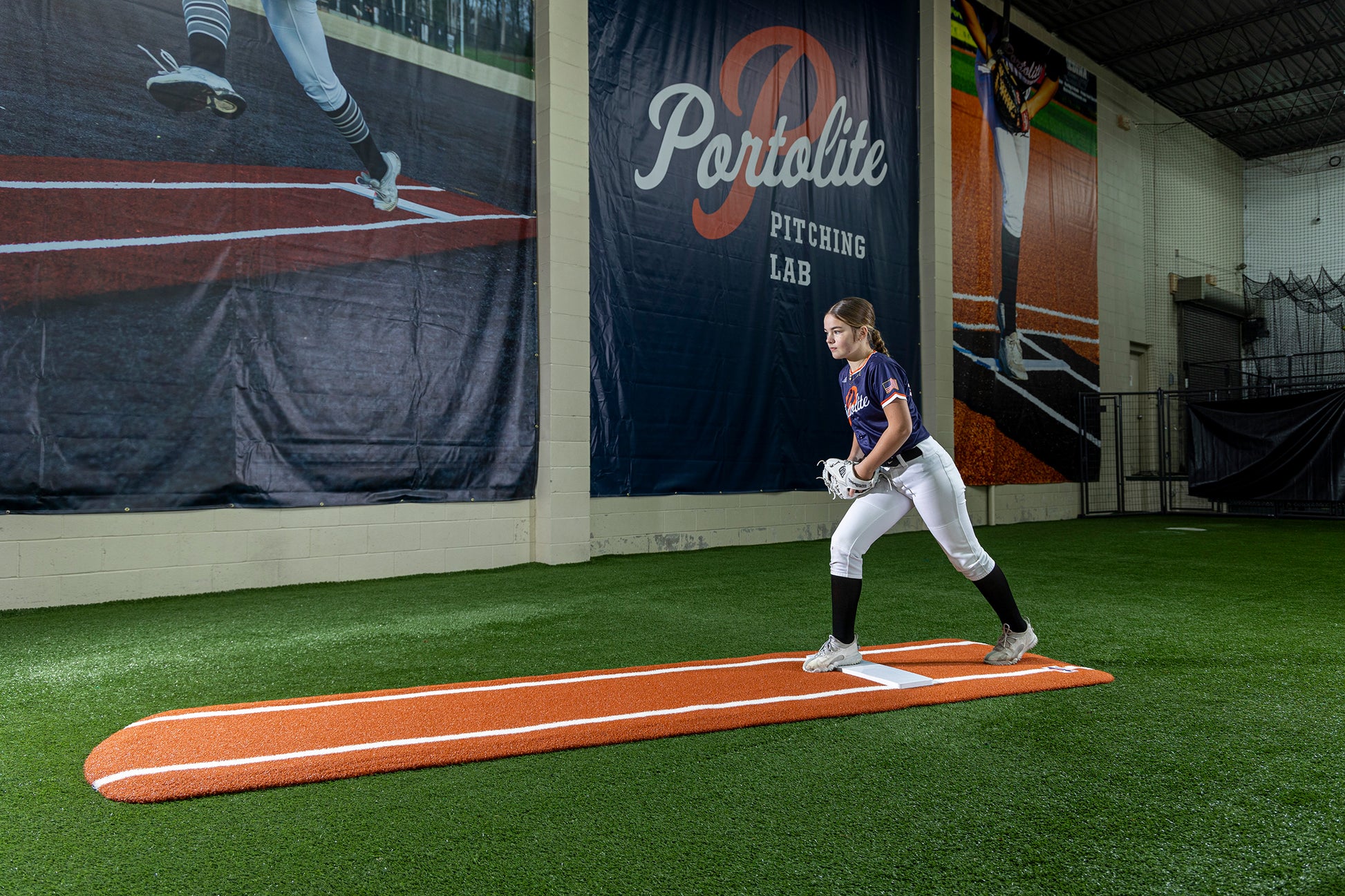 A softball player stands ready to pitch on a Portolite Signature Practice Mat with Spikes with Lines / Lanes, built for 12U pitchers, in an indoor facility with synthetic turf and a Portolite Pitching Lab banner displayed prominently behind her.
