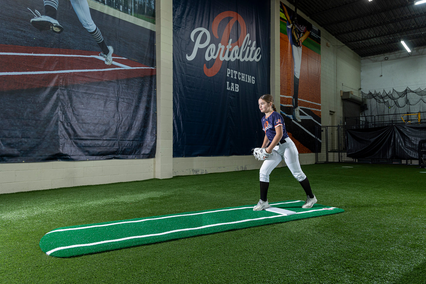 A young softball player in uniform stands on a Portolite Signature Practice Mat with Spikes with Lines / Lanes—built for 12U pitchers—inside an indoor facility with synthetic turf, holding a glove, with a Portolite Pitching Lab banner on the wall behind her, ready to perfect her pitching drills.