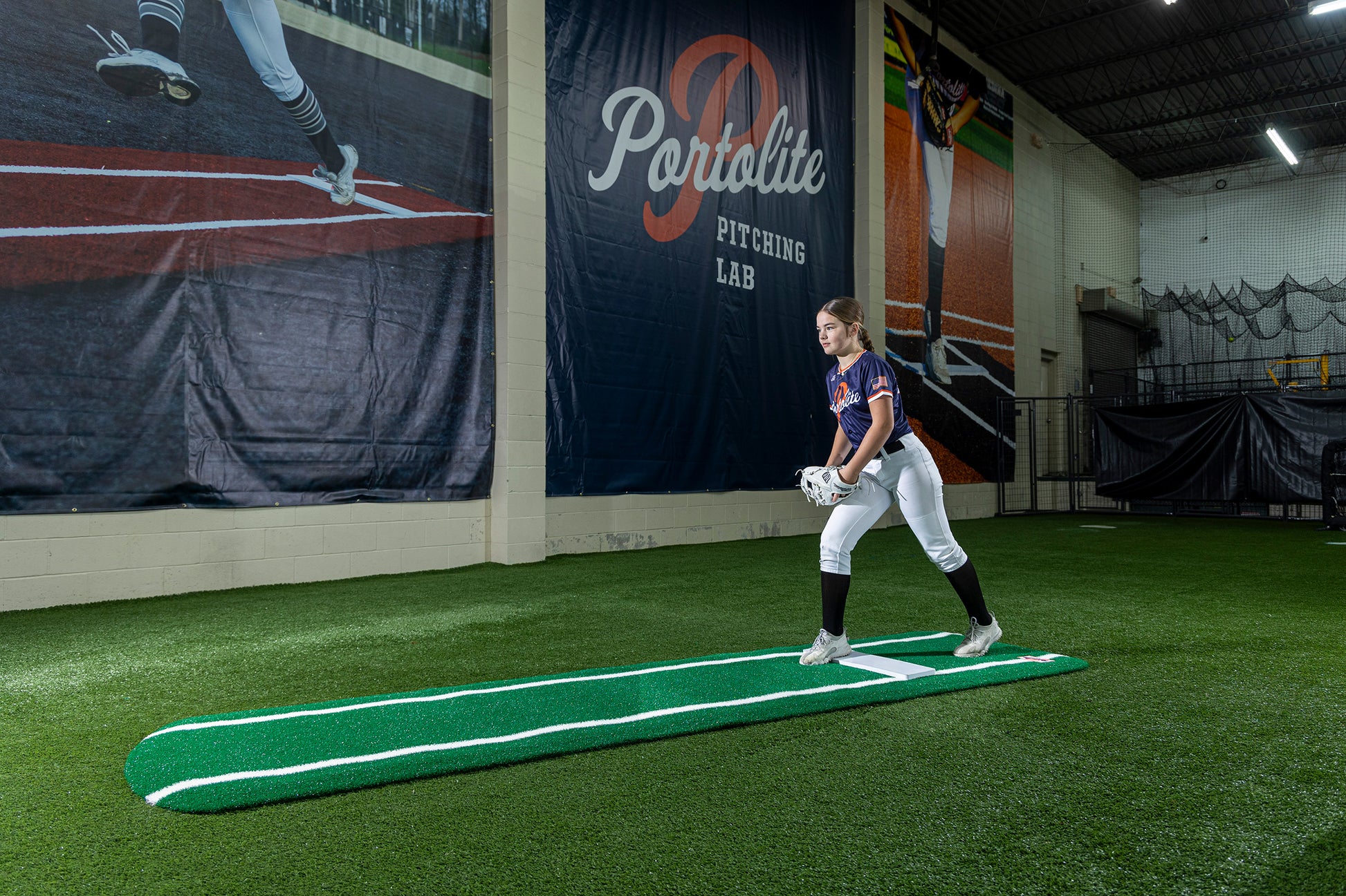 A young softball player in uniform stands on a Portolite Signature Practice Mat with Spikes with Lines / Lanes—built for 12U pitchers—inside an indoor facility with synthetic turf, holding a glove, with a Portolite Pitching Lab banner on the wall behind her, ready to perfect her pitching drills.