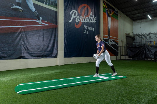 A young softball player in uniform stands on a Portolite Signature Practice Mat with Spikes with Lines / Lanes—built for 12U pitchers—inside an indoor facility with synthetic turf, holding a glove, with a Portolite Pitching Lab banner on the wall behind her, ready to perfect her pitching drills.