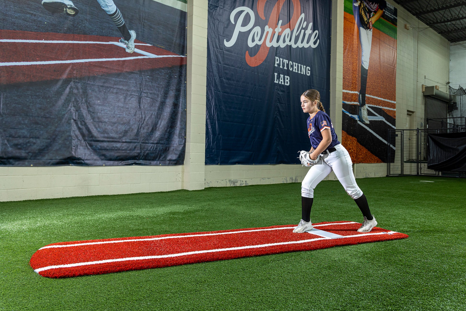 A young softball player stands indoors on the Portolite Signature Practice Mat with Spikes with Lines / Lanes, built for 12U pitchers, preparing to pitch. Large posters of players and the Portolite Pitching Lab are visible on the wall behind her, while the synthetic turf beneath her feet offers an ideal surface for pitching drills.