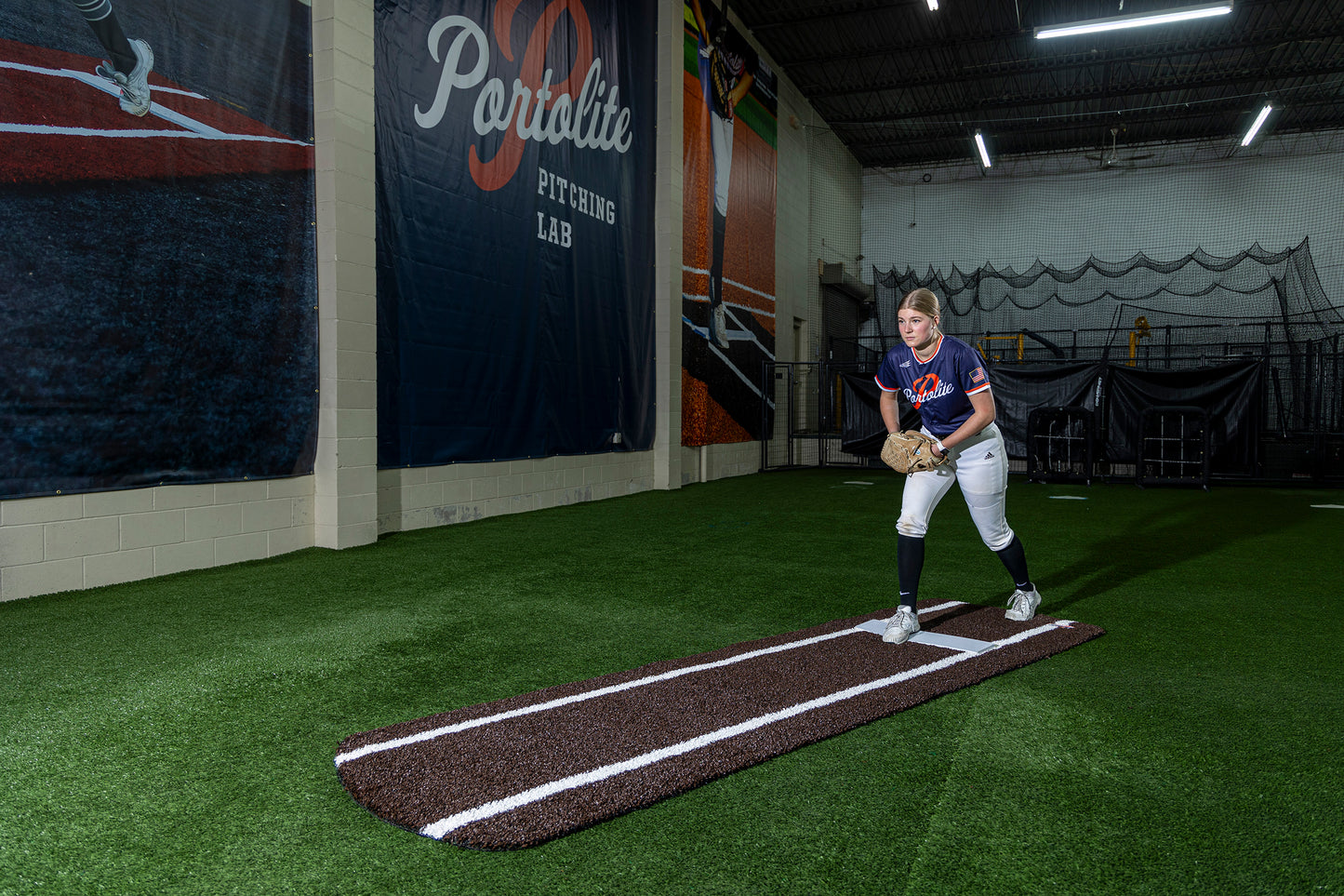 A softball pitcher wearing a Portolite jersey stands poised on the green turf at the Portolite Pitching Lab, preparing for bullpen practice on the Portolite Ultimate Spiked Practice Mat with Lanes and Lines, a premium mat built for all ages and skill levels.