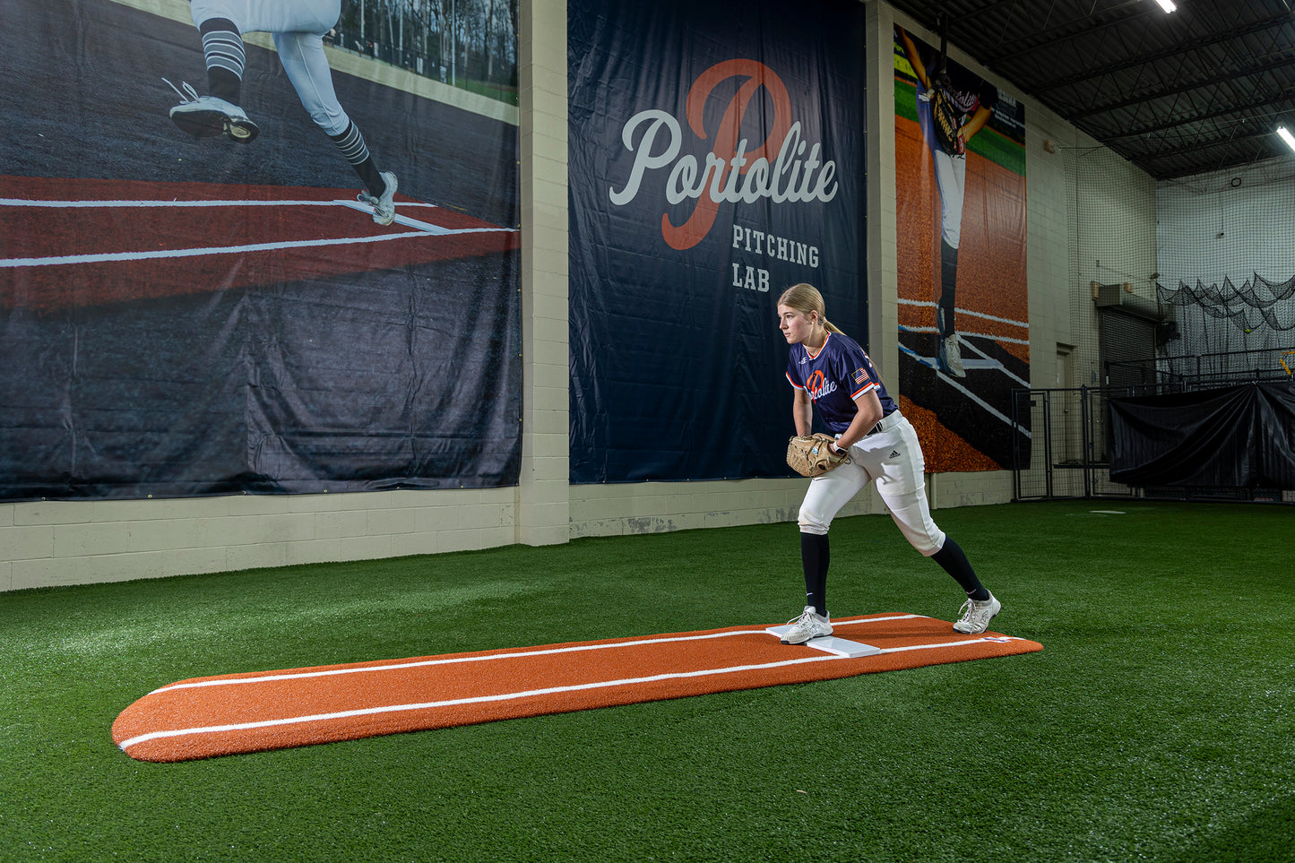A softball player stands on the Portolite Ultimate Spiked Practice Mat with Lanes / Lines indoors, preparing to throw. Behind her is a large mural featuring a pitcher and the words “Portolite Pitching Lab,” creating the perfect environment for focused bullpen practice. Built for all ages and skill levels.