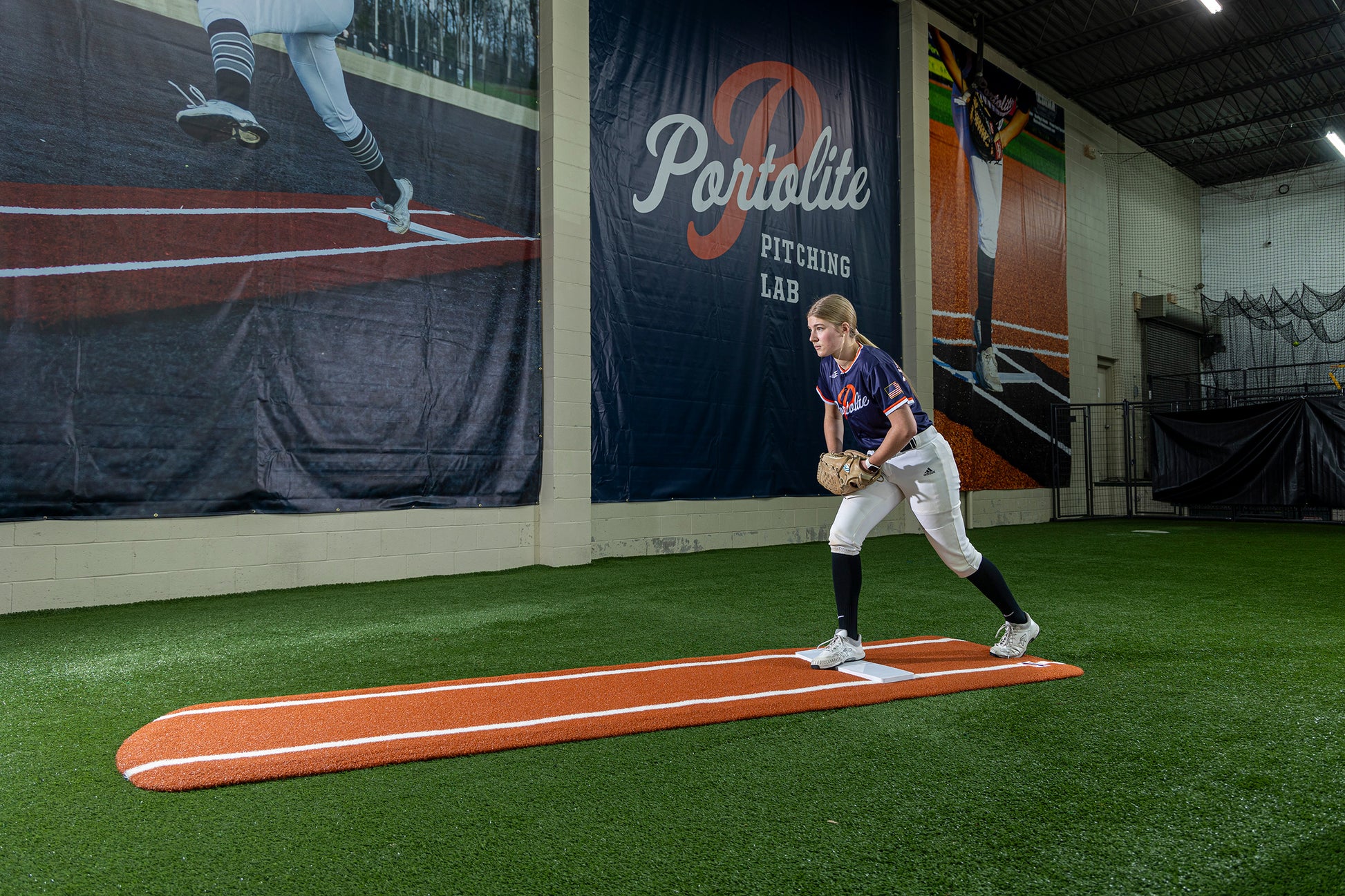 A softball player stands on the Portolite Ultimate Spiked Practice Mat with Lanes / Lines indoors, preparing to throw. Behind her is a large mural featuring a pitcher and the words “Portolite Pitching Lab,” creating the perfect environment for focused bullpen practice. Built for all ages and skill levels.