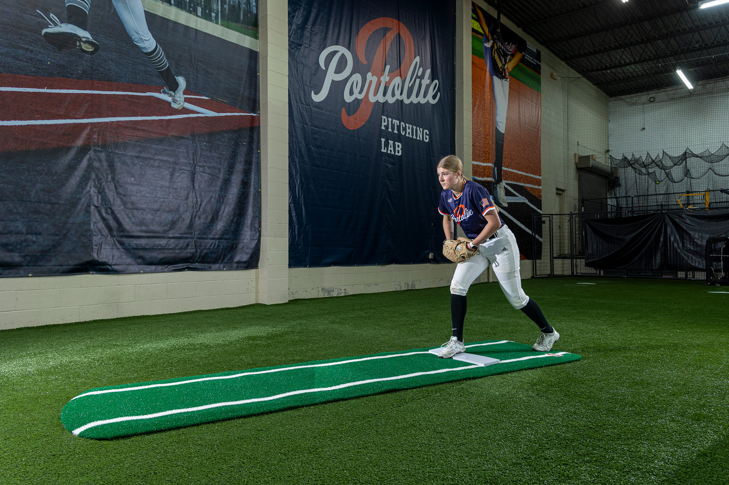 At the Pitching Lab facility, a softball player stands on the Portolite Ultimate Spiked Practice Mat with Lanes / Lines—built for all ages and skill levels—wearing a glove and preparing to pitch, creating an ideal environment for focused bullpen practice.