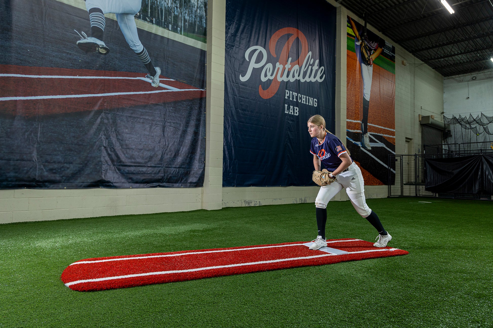 A softball player stands on a Portolite Ultimate Spiked Practice Mat with Lanes / Lines inside an indoor facility, preparing to pitch. A large Portolite Pitching Lab banner hangs on the wall behind her, creating an ideal setting for bullpen practice. The mat is built for all ages and skill levels.