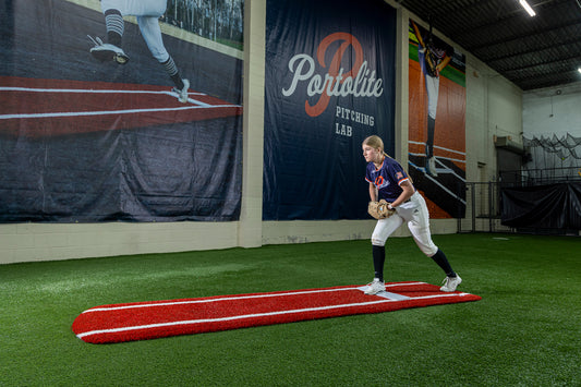 A softball player stands on a Portolite Ultimate Spiked Practice Mat with Lanes / Lines inside an indoor facility, preparing to pitch. A large Portolite Pitching Lab banner hangs on the wall behind her, creating an ideal setting for bullpen practice. The mat is built for all ages and skill levels.