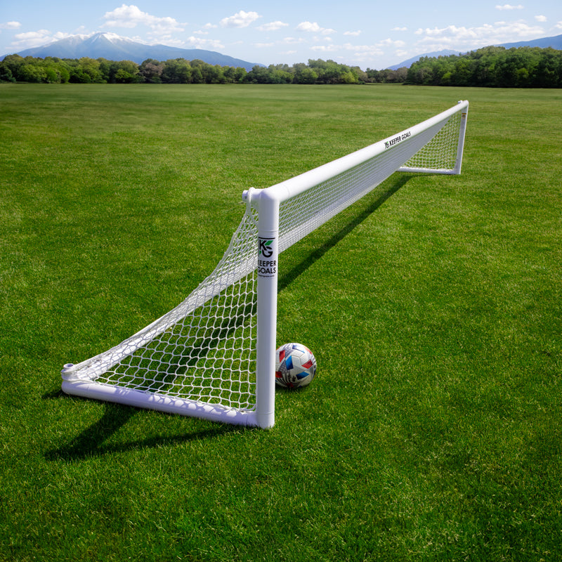 A Precizion Soccer Goal net spans a grassy field with a soccer ball nearby. Trees and distant mountains sit under a blue, cloud-scattered sky—ideal for practicing accuracy using quality soccer training gear.