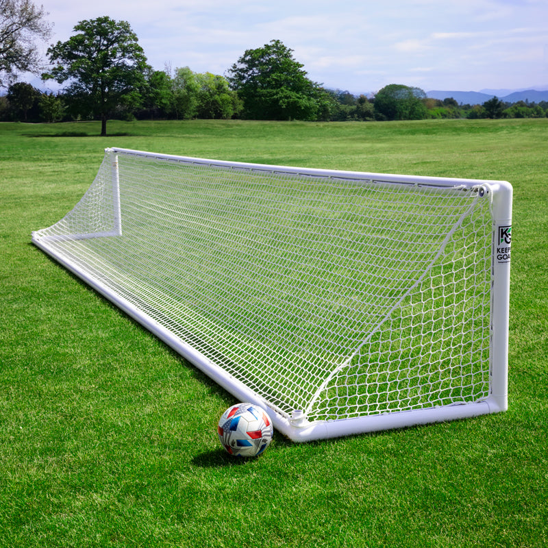A Precizion Soccer Goal with a durable net stands on a grassy field under clear skies; perfect for accuracy, a soccer ball rests by the goalpost. Trees and rolling hills form the backdrop.