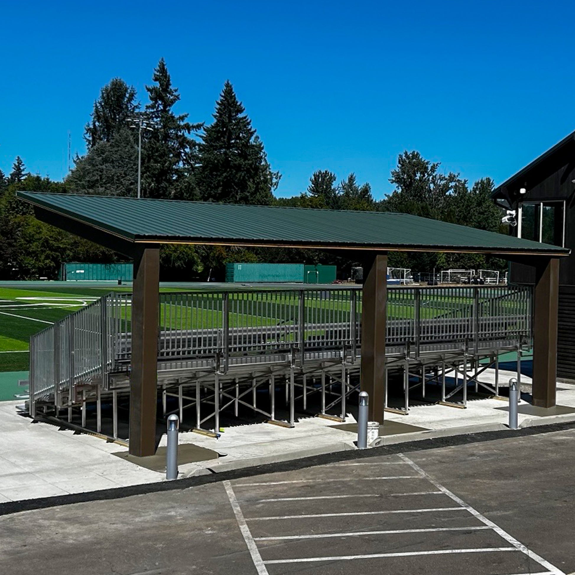 Bleachers with the Premium Hard Shell Bleacher Cover are near an artificial turf sports field. Trees and a blue sky appear in the background, and an empty parking lot is visible in the foreground.