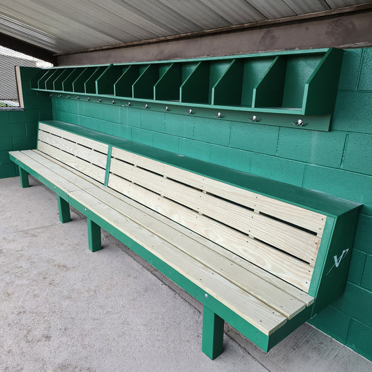 The Condor Baseball & Softball Dugout Bench is a two-tiered wood player bench with cubby storage and coat hooks, mounted on a green cinderblock wall under a metal roof—ideal for baseball and softball programs. The bench and cubbies are empty.