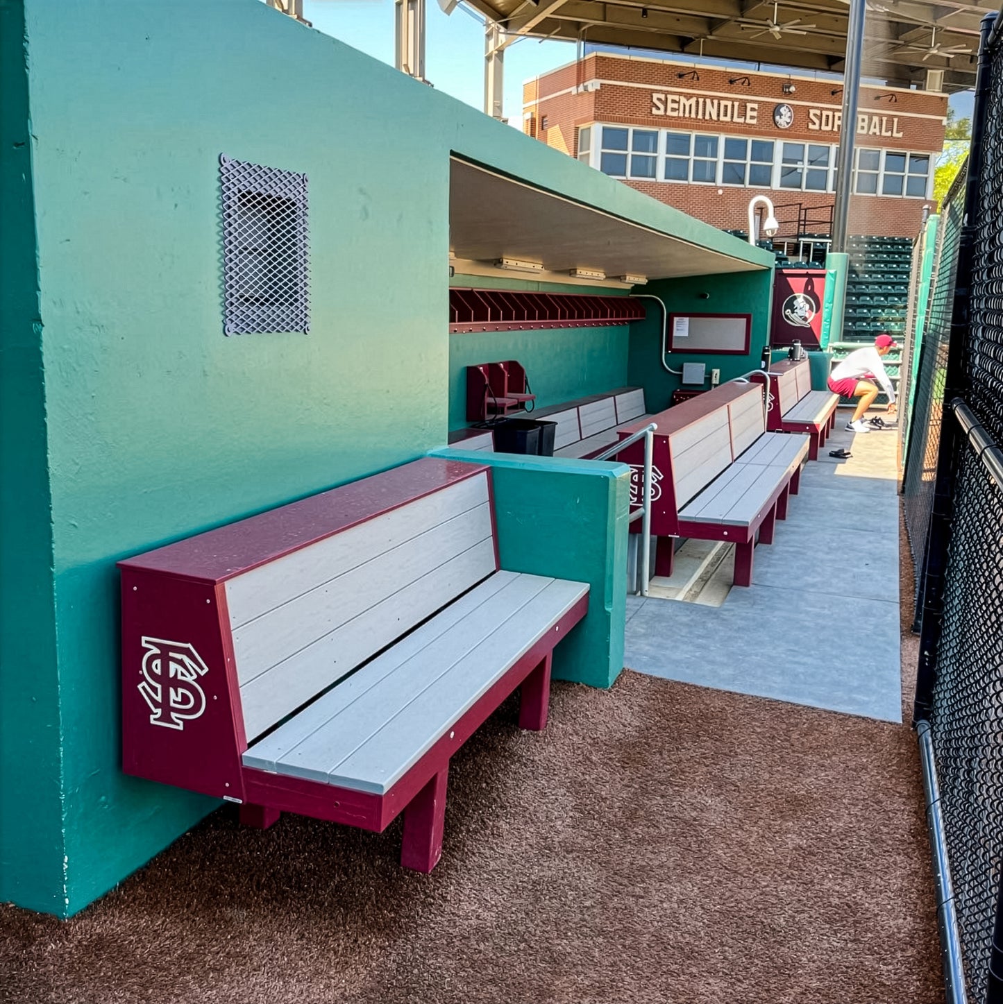 A softball dugout featuring Eagle Dugout Bench MLB-Style Wood Benches in maroon and gray, teal walls, and sports gear. The Seminole Softball stadium and a uniformed player appear in the background.