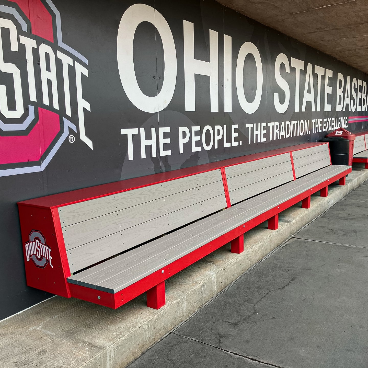 The Eagle Dugout Bench | MLB-Style Wood Baseball & Softball Bench with red trim sits by a wall displaying large OHIO STATE BASEBALL text and the Ohio State logo, ideal for any stadium's dugout area.