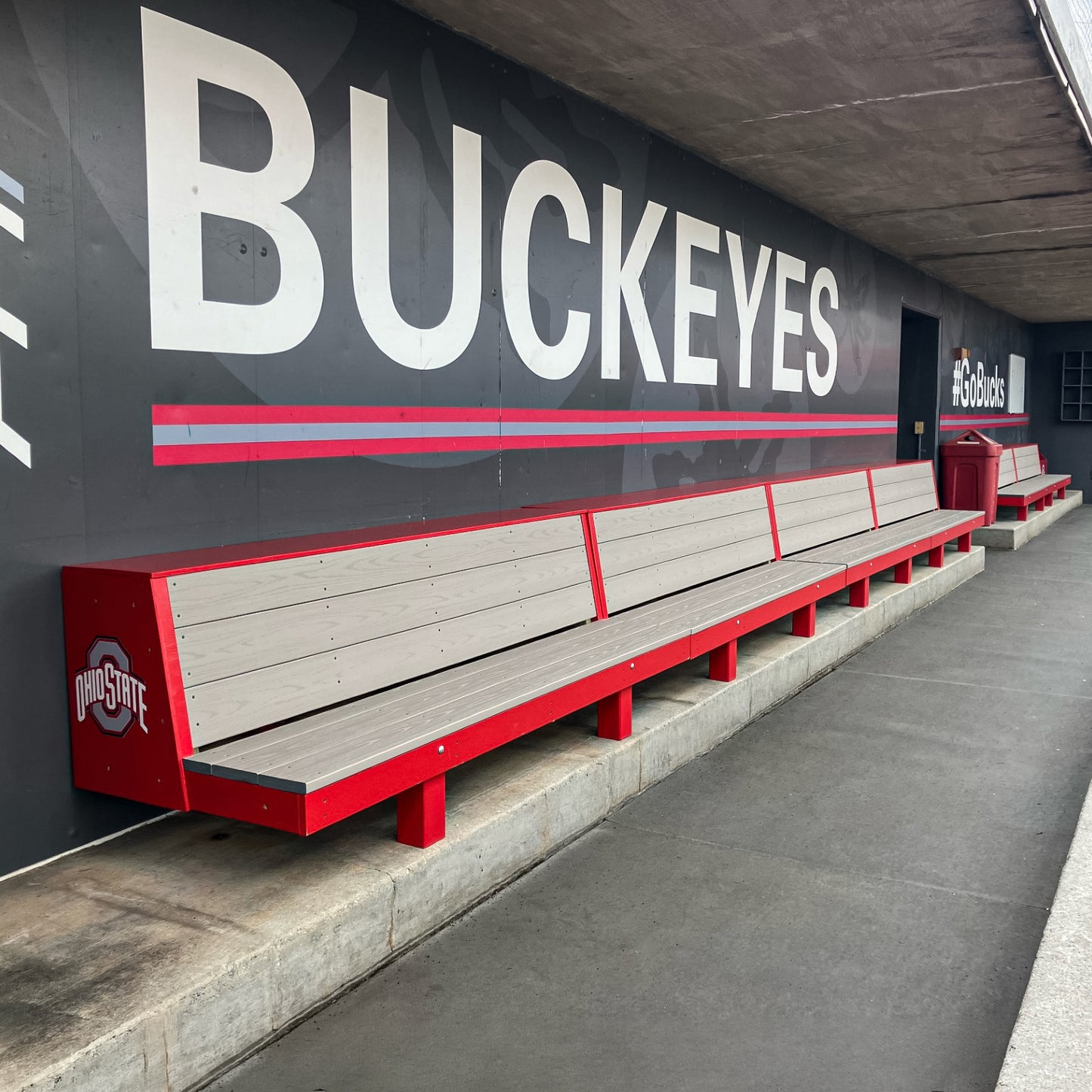 The Eagle Dugout Bench | MLB-Style Wood Baseball & Softball Bench, in red and gray with the Ohio State logo, sits by a concrete wall displaying large BUCKEYES letters and #GoBucks signage at an Ohio State sports facility.