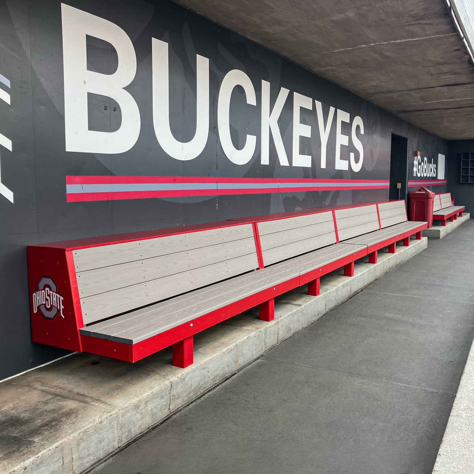 The Eagle Dugout Bench | MLB-Style Wood Baseball & Softball Bench, in red and gray with the Ohio State logo, sits by a concrete wall displaying large BUCKEYES letters and #GoBucks signage at an Ohio State sports facility.