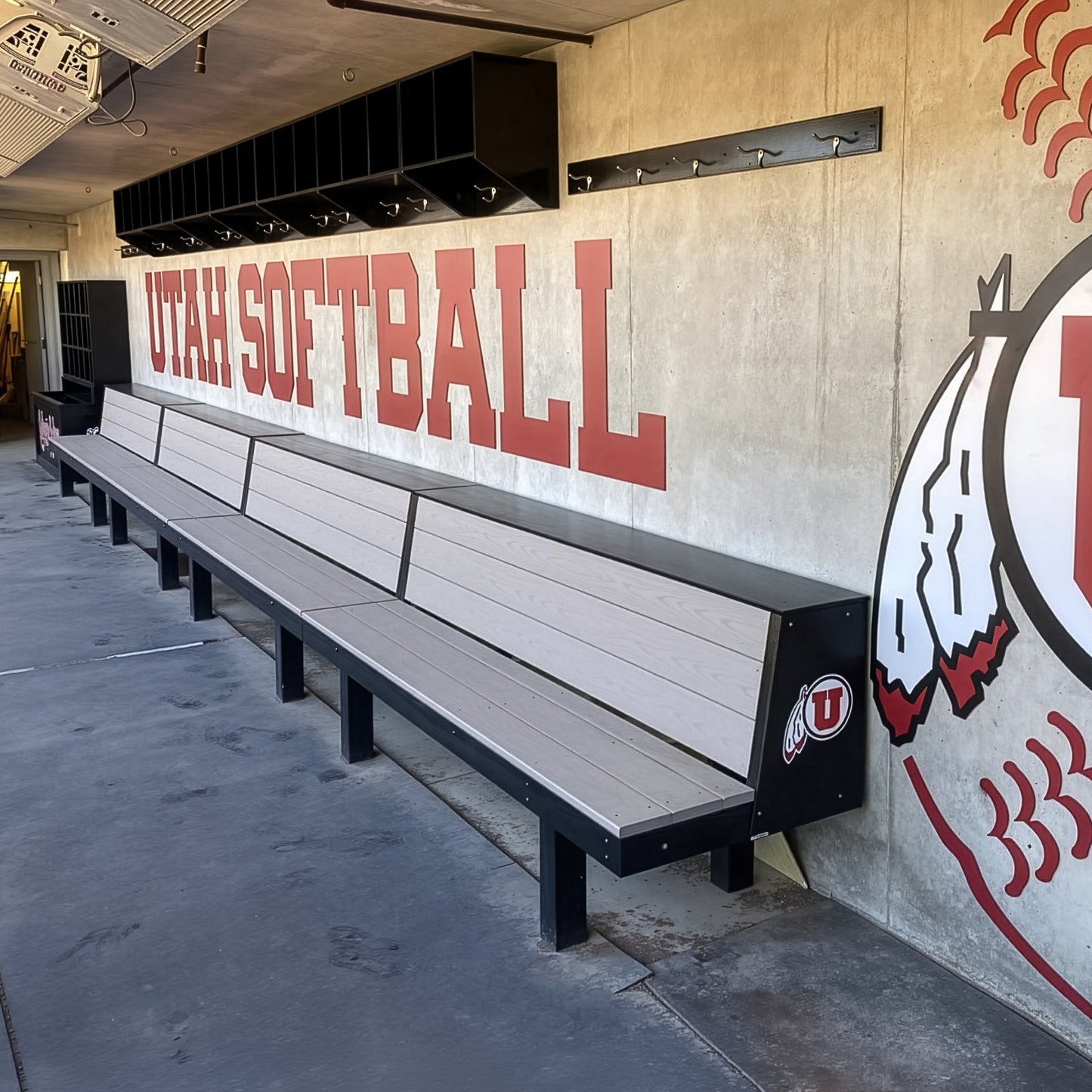 A dugout featuring the Eagle Dugout Bench | MLB-Style Wood Baseball & Softball Bench, with cubby storage above and wall hooks. "Utah Softball" and a large University of Utah logo are painted on the concrete wall.