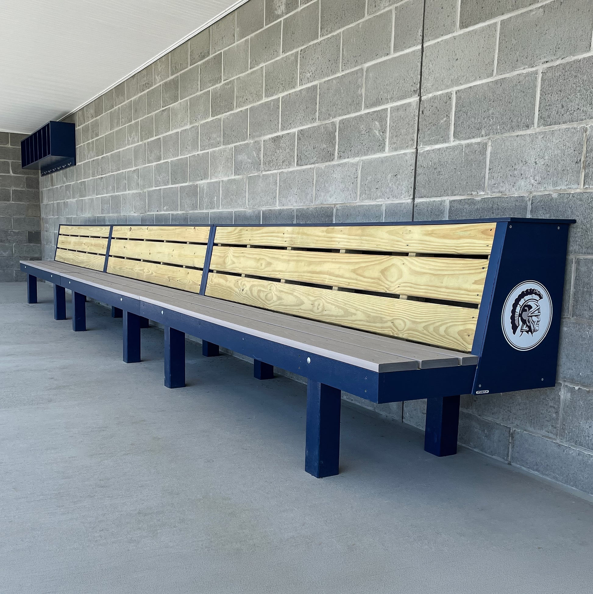 The Falcon Dugout Bench | MLB-Style 2-Tier Wood Baseball & Softball Bench features a wooden and blue metal design with a Spartan helmet logo, perfect for outdoor team areas or dugouts against concrete block walls.