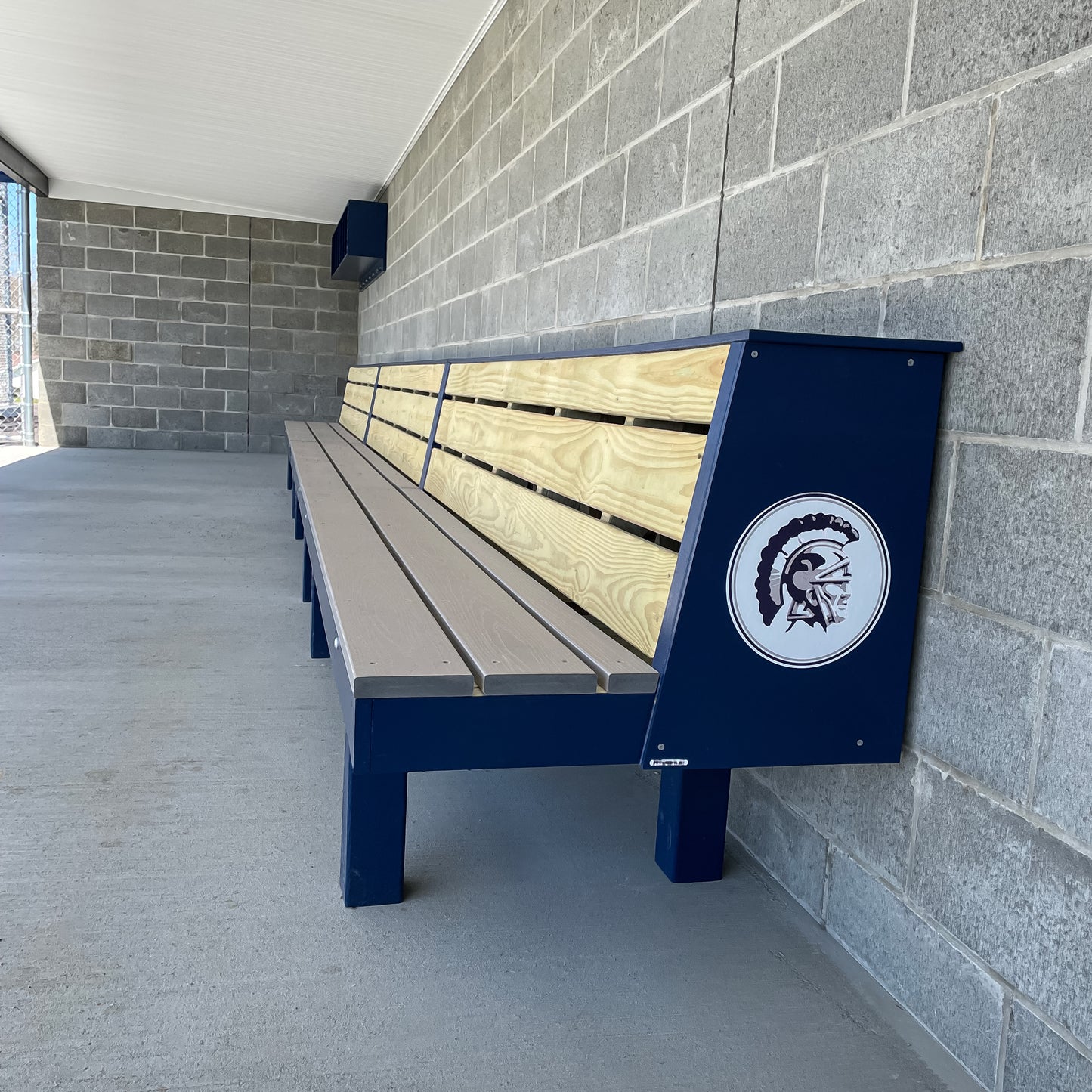The Falcon Dugout Bench, an MLB-style 2-tier wood baseball & softball bench with blue metal supports, sits against a cinder block wall. Its end displays a Spartan helmet logo. The area appears clean and well-lit.