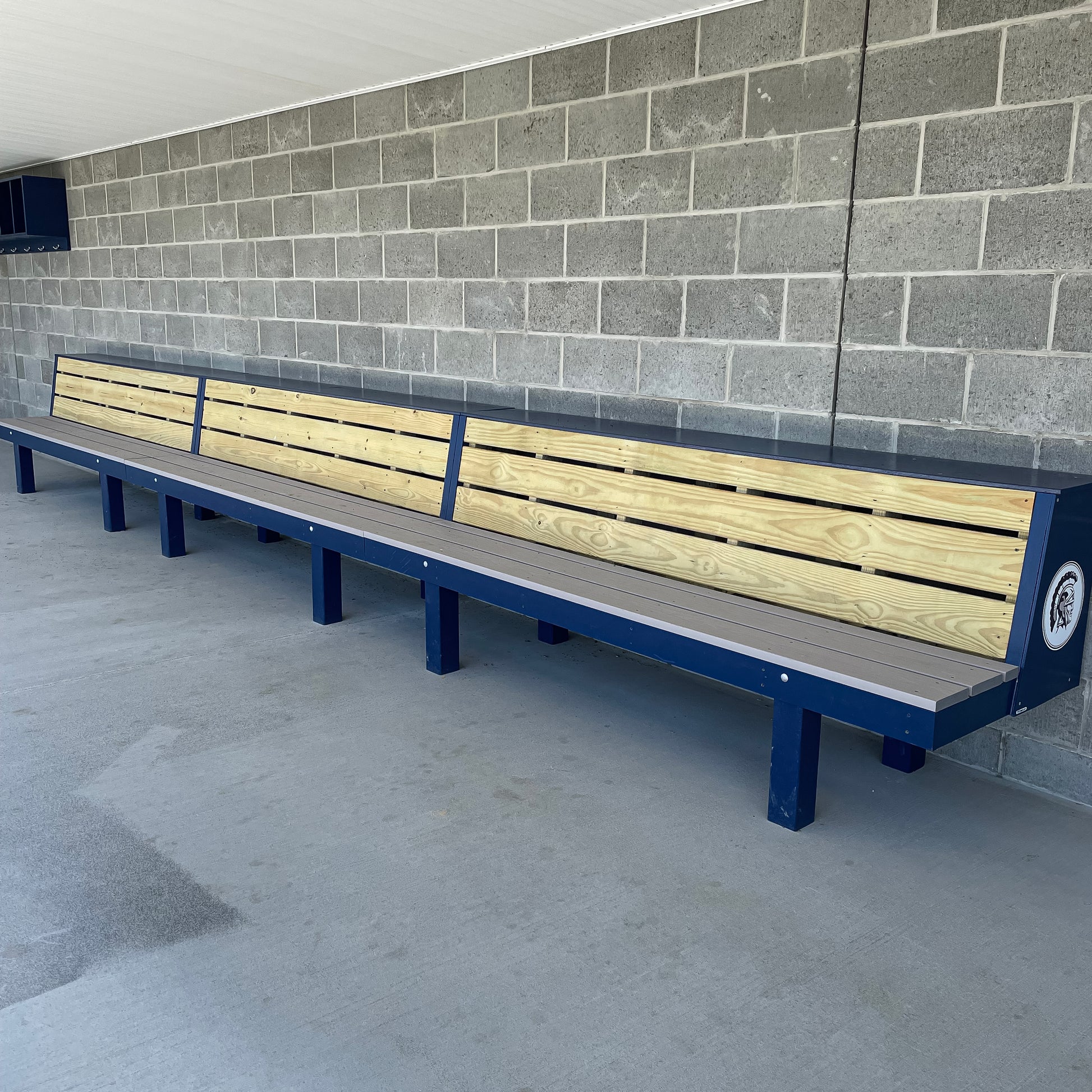 The Falcon Dugout Bench, an MLB-style 2-tier wood baseball & softball bench with a blue metal frame, sits against a gray cinder block wall on concrete, creating an authentic dugout look in your sports facility.