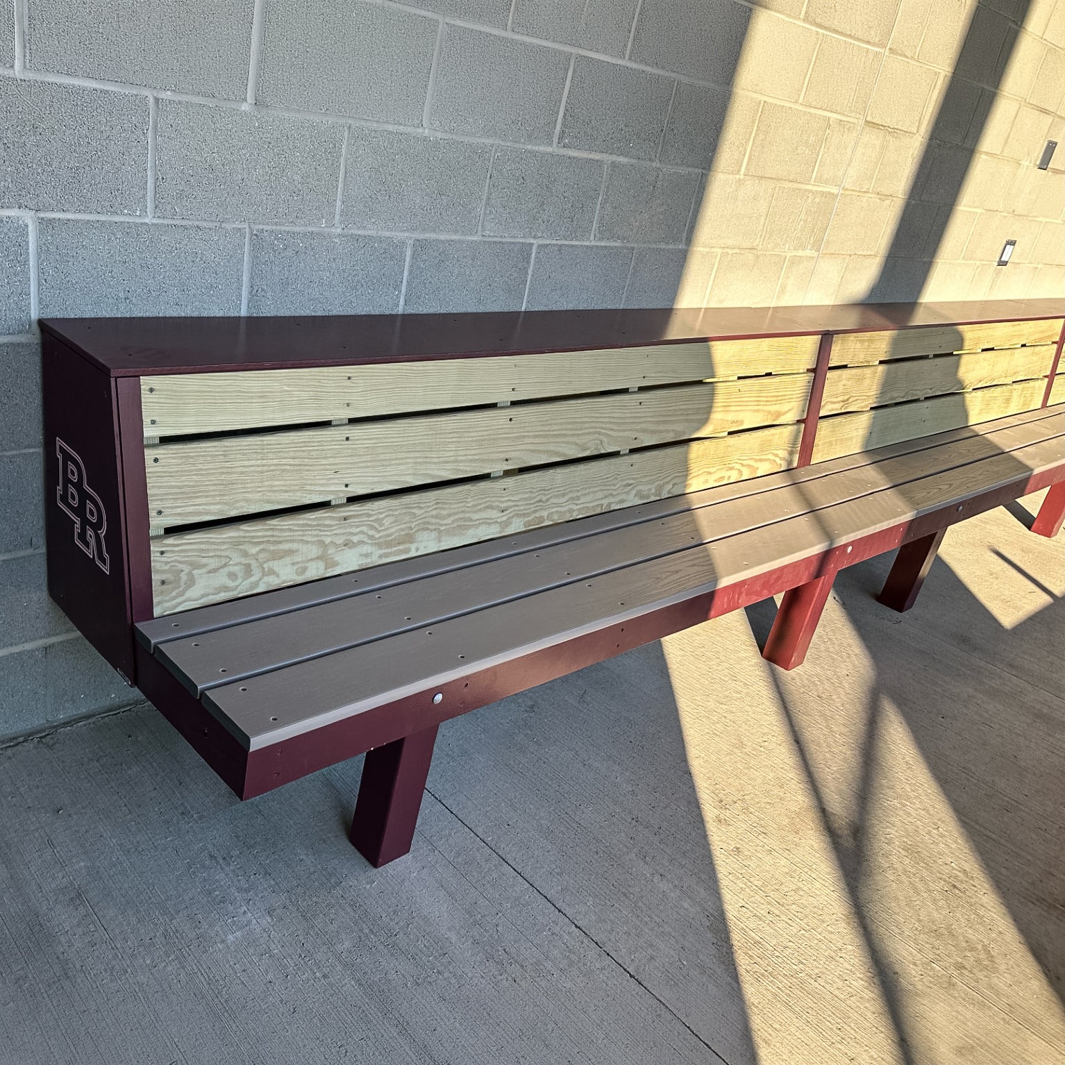 A Falcon Dugout Bench | MLB-Style 2-Tier Wood Baseball & Softball Bench with a maroon metal frame and BR initials sits against a light brick wall, casting shadows from the bench and passersby onto the ground and wall.