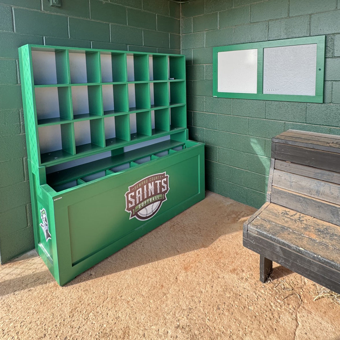 The Field General Dugout Helmet and Bat Rack is an 18-helmet baseball & softball storage unit, perfect for dugouts with green brick walls, a wooden bench, gray boards, and a dirt floor to organize helmets and bats with the Saints Baseball logo.