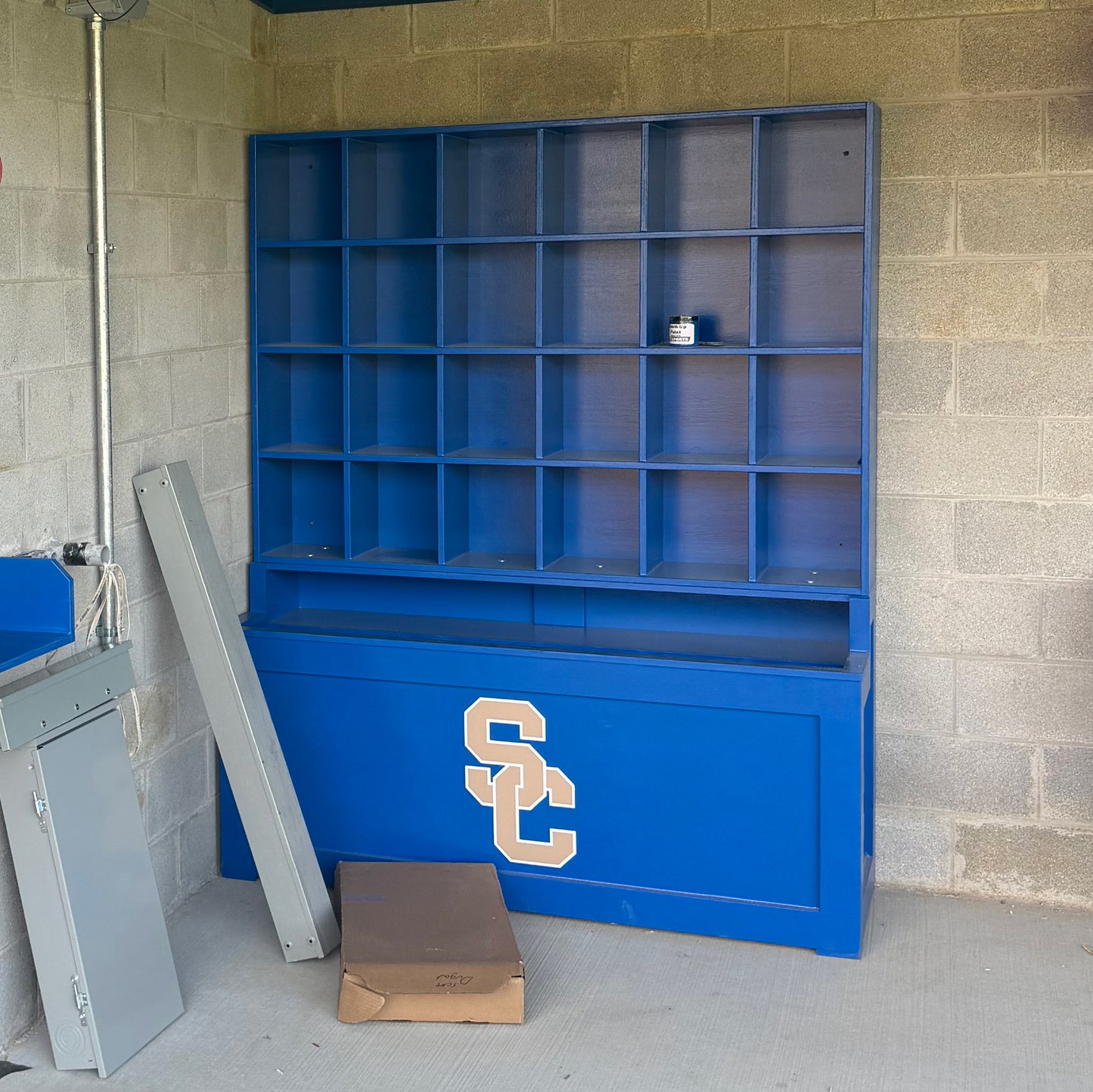 A Field Marshall Dugout Helmet and Bat Rack | 24-Helmet Baseball & Softball Storage Unit with SC on the front stands against a cinder block wall. A can sits on a shelf near a cardboard box and two gray metal panels leaning nearby.