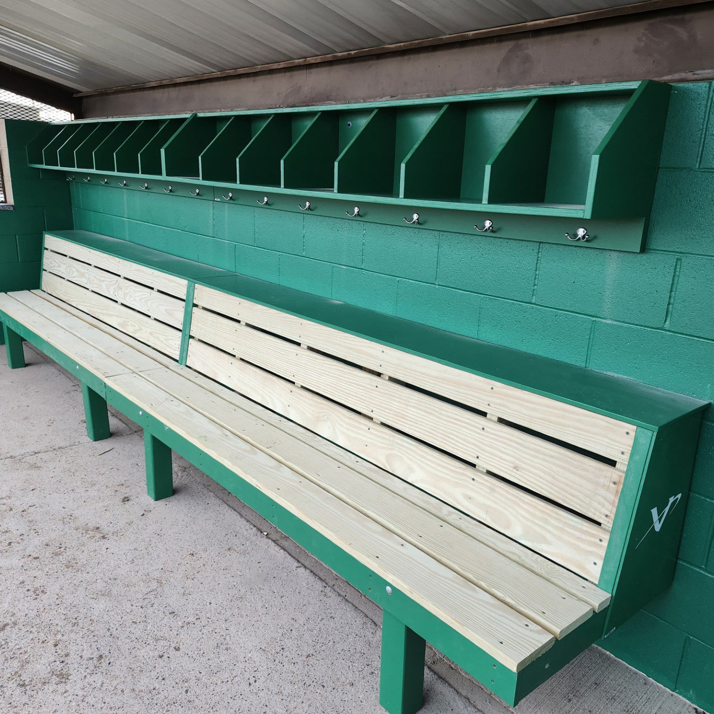 The Geary Dugout Storage Rack is a green and light wood organizer with open-top cubbies and hooks, mounted above a bench against a green cinder block wall under a metal roof in the dugout.