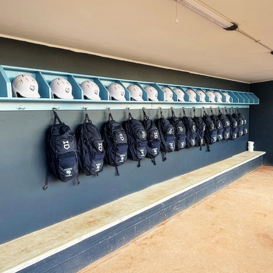 White baseball helmets sit on the Geary Dugout Storage Rack above black backpacks in a clean dugout, featuring a concrete bench and dirt floor—ideal for organizing gear in baseball and softball team settings.