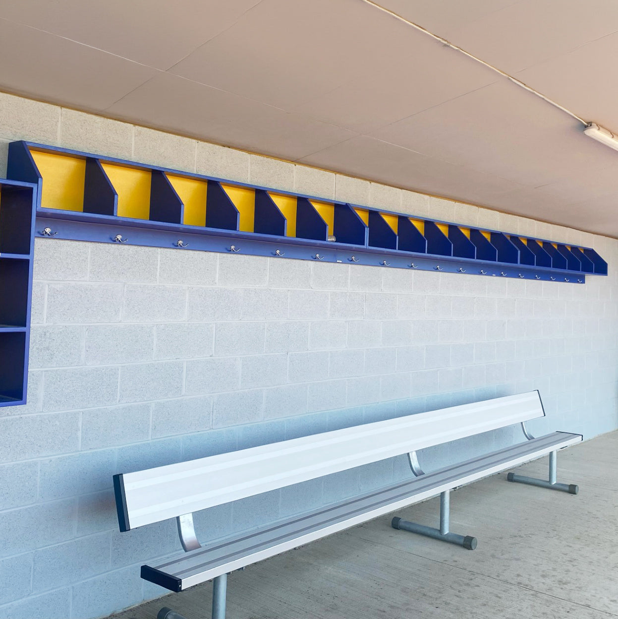 The Geary Dugout Storage Rack, an above-bench baseball and softball dugout organizer with open-top cubbies, is mounted on a gray cinder block wall above a metal bench. Blue and yellow accents complete the dugout storage area.