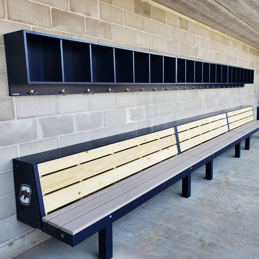 A Premier Dugout Storage Rack | Baseball and Softball Dugout Organizer with open lockers and hooks runs above a long wooden bench with a backrest against a concrete dugout wall, featuring a sports team logo on one end.