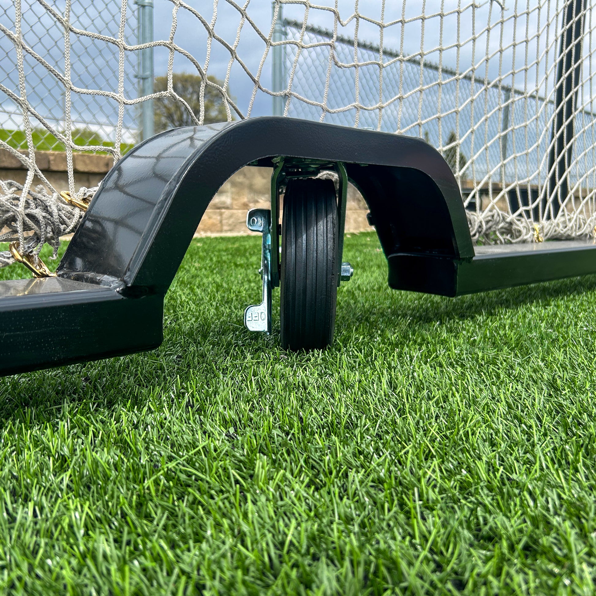 A close-up of the wheel attachment on the Ultimate Wheeled Box-Style Soccer Goal positioned on artificial turf, showing the wheel beneath the frame with the net and a portion of the field visible in the background.