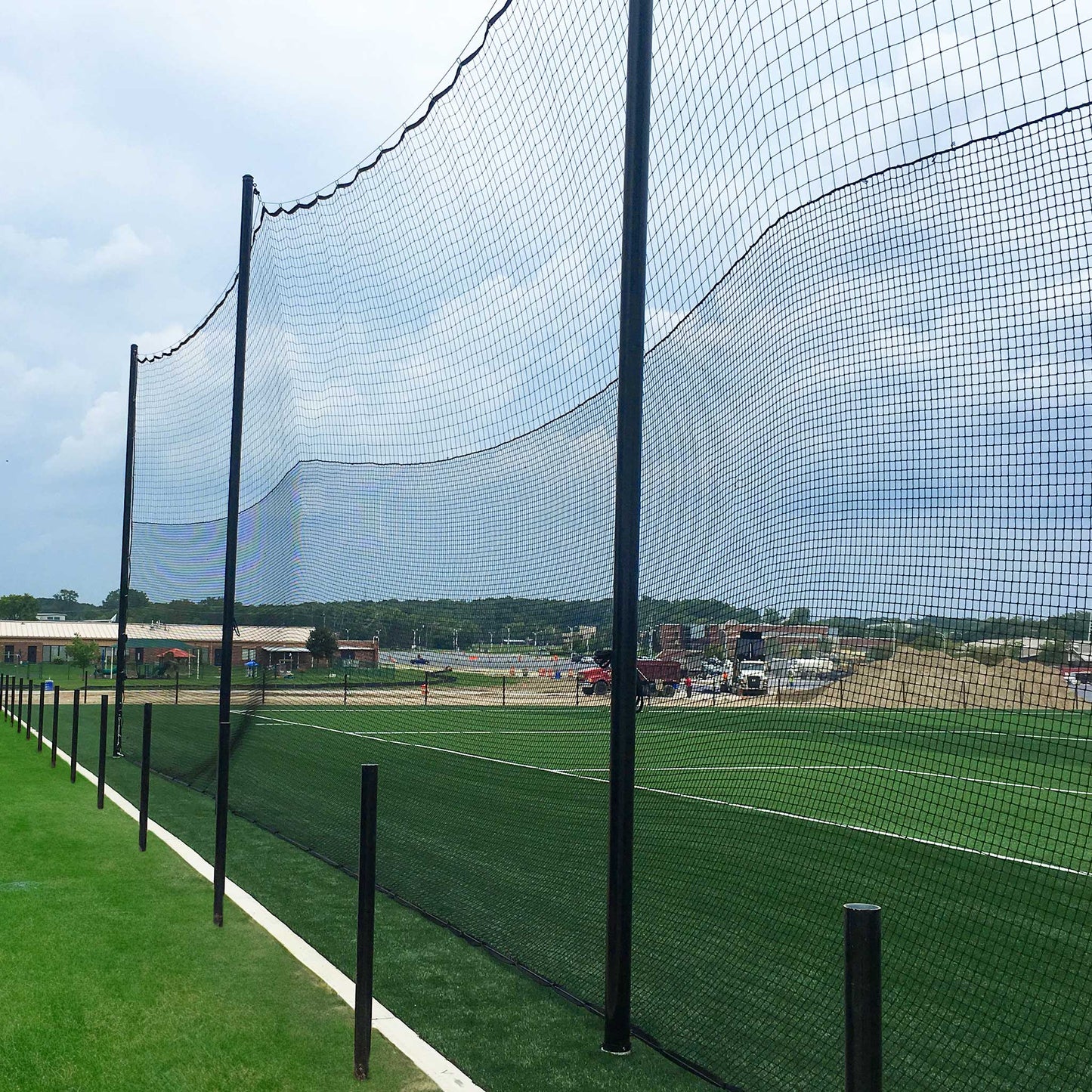 Hybrid Soccer & Lacrosse Backup Netting Systems with tall black netting and sturdy poles border a green artificial turf field, with buildings, vehicles, and a sandy construction area visible in the cloudy background.