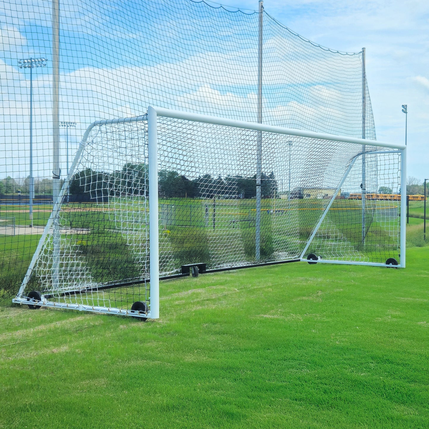 An 8 x 24 striped soccer net (4mm PE, 4T x 8B) stands on green grass at a sports field, backed by larger nets and under a partly cloudy blue sky.