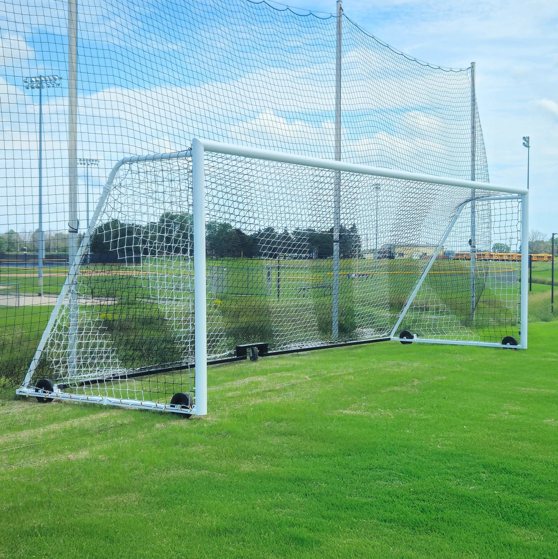 An 8 x 24 striped soccer net (4mm PE, 4T x 8B) stands on green grass at a sports field, backed by larger nets and under a partly cloudy blue sky.
