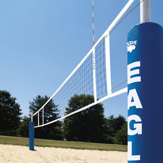 A Premium Competition Sand Volleyball System with a white net and blue padded poles—one labeled EAGLE—stands on sand amid green grass, trees, and under a clear blue sky.