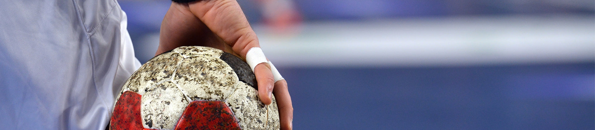 A close-up of a person’s hand holding a worn handball, with fingers taped for support. The background is blurred, drawing focus to the ball and hand.