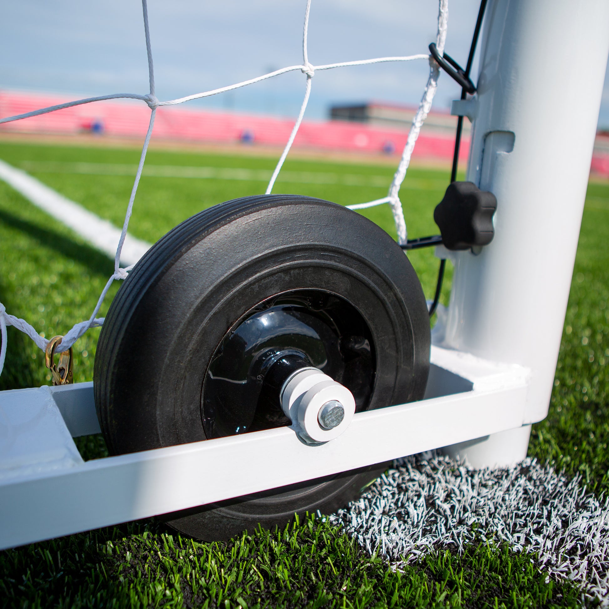 Close-up of the Ultimate Wheeled Box-Style Soccer Goals black wheel on artificial turf near a white boundary line, ensuring easy movability. Features netting and a professional stadium backdrop for an authentic playing experience.