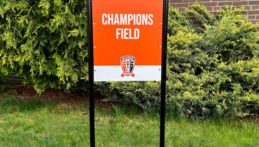 Custom field sign for sports field. Black u-shaped frame on circular base. Orange and white sign with words Champions Field and logo.