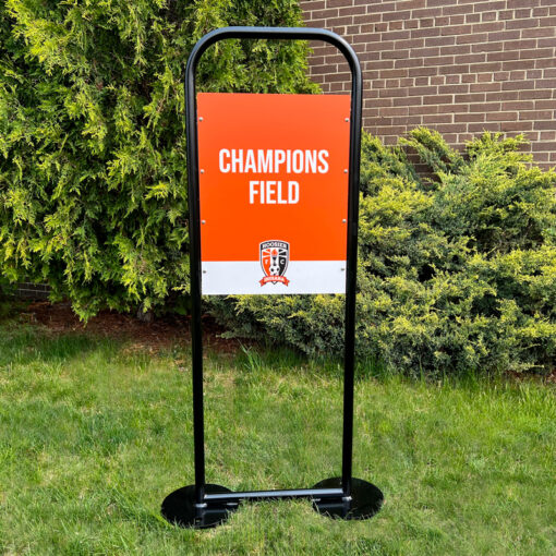 Custom field sign for sports field. Black u-shaped frame on circular base. Orange and white sign with words Champions Field and logo.