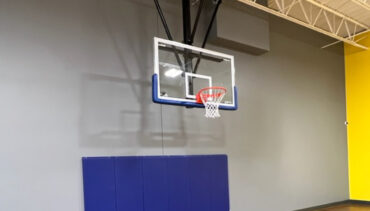 Ceiling mounted basketball hoop in rec center gym. Blue padding on gray wall behind the hoop.