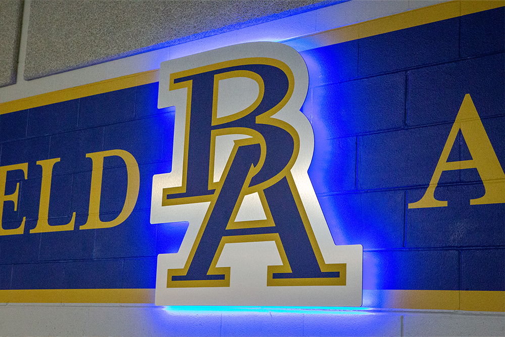 Royal blue logo of the letters B and A outlined in yellow on a band of royal blue with yellow border on a brick wall.